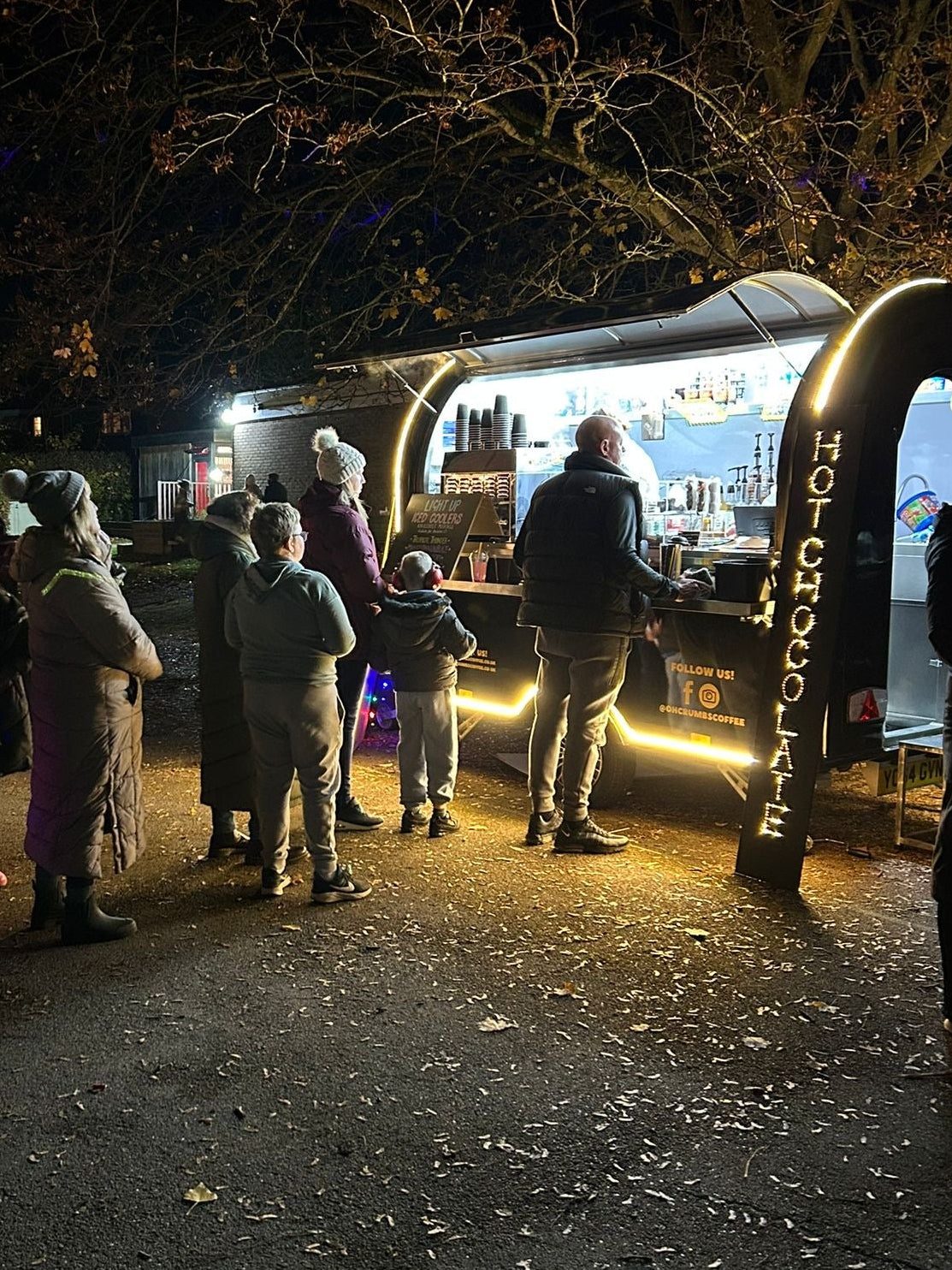 People gathered around an Oh Crumbs! Coffee trailer at night with warm lighting.